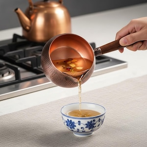 May include: A hammered copper pouring vessel with a dark wooden handle, featuring a built-in strainer, pouring a light-colored tea into a small white ceramic bowl decorated with blue floral patterns, with a copper kettle visible in the background.