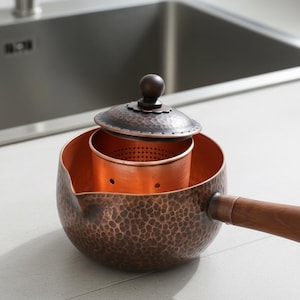 May include: A hammered copper teapot with a dark brown wooden handle and a matching lid. The interior features a removable copper tea infuser basket with small perforations. The exterior has a textured, dimpled finish and a small pouring spout.