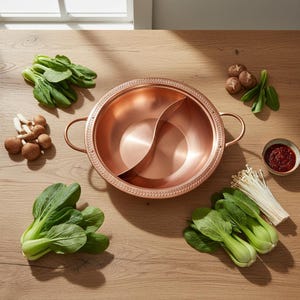Puede incluir: Una olla redonda de color cobre con dos asas laterales y un divisor central. El borde presenta un patrón texturizado. Está rodeada de bok choy fresco, racimos de champiñones marrones, un cuenco de salsa roja y setas enoki blancas.