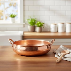 May include: A round, hammered copper pan with two side handles, positioned on a wooden surface next to a light-colored cloth napkin and a wooden spoon. The pan features a metallic, reddish-brown finish with a textured, dimpled surface pattern.