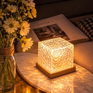 May include: A square glass lamp with a textured, rippled surface that emits a warm, golden light. The lamp sits on a flat, square wooden base, casting intricate, water-like light patterns onto the surface below it.