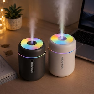 以下が含まれることがあります： マットブラックとオフホワイトの円筒形加湿器が2台並んでおり、それぞれに「HUMIDIFIER」の文字が縦書きされています。どちらのユニットも、虹色に光る円形のトップパネルから垂直にミストを噴出しています。