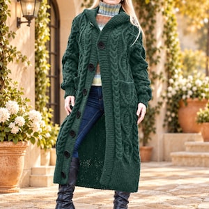 May include: A long, dark green cable-knit cardigan featuring a hood, long sleeves, and a front button closure. The garment is worn over a multi-colored striped mock-neck sweater and dark blue denim jeans, paired with black knee-high boots.
