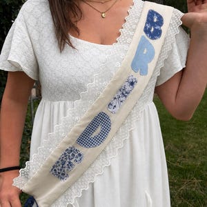 May include: A cream-colored sash featuring white lace trim along the edges and the word BRIDE spelled out in large, fabric-appliqued letters. The letters feature various blue patterns, including floral prints, gingham checks, and solid light blue.
