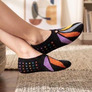 May include: A pair of black crochet slippers featuring a colorful fan-shaped pattern on the toe in yellow, orange, and purple, with a grid of small multi-colored cross-stitch accents along the sides and a low-cut opening at the top.