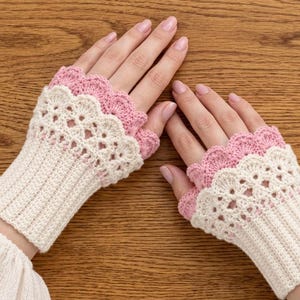Könnte beinhalten: Ein Paar fingerlose Handschuhe mit einem cremefarbenen gerippten Bündchen und einem zarten weißen Häkelspitzenmuster. Der obere Rand jedes Handschuhs ist mit einem gewellten rosa Saum versehen, was für eine geschichtete Optik an der Hand sorgt.