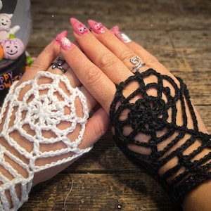 May include: A pair of fingerless gloves featuring a crocheted spider web pattern, with one glove in white and the other in black. The design covers the back of the hand and wrist, leaving the fingers exposed, and is crafted from yarn.