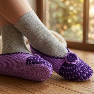 Può includere: Un paio di pantofole all'uncinetto viola con un motivo a maglia testurizzato, una sezione della punta viola scuro e un piccolo fiore decorativo all'uncinetto sulla parte superiore di ogni pantofola, indossate sopra calzini in maglia grigio chiaro.