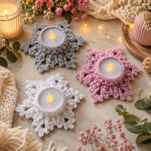 May include: Three crochet candle holders in gray, pink, and white, each shaped like a six-pointed star. Each holder features a circular center containing a small, round, battery-operated LED tea light with a glowing yellow flame.