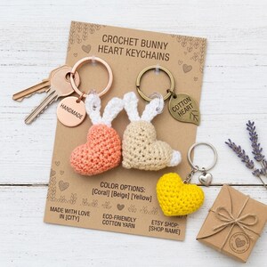 May include: Three crochet heart keychains in coral, beige, and yellow on a display card labelled CROCHET BUNNY HEART KEYCHAINS. The coral and beige hearts feature white bunny ears. Includes metal key rings, a HANDMADE tag, and a COTTON HEART leaf charm.