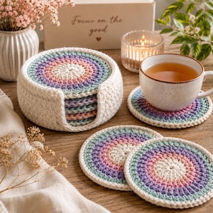 May include: A set of round, crocheted coasters featuring a gradient of pastel colors including pink, purple, and light blue, with a cream-colored border. The coasters are stored in a matching cream-colored, circular crocheted holder.