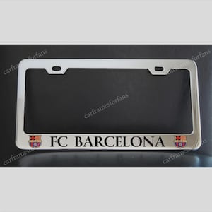 May include: A rectangular chrome licence plate frame featuring the text FC BARCELONA in black lettering centred at the bottom. The frame includes two small FC Barcelona team crests on either side of the text and two mounting holes at the top.