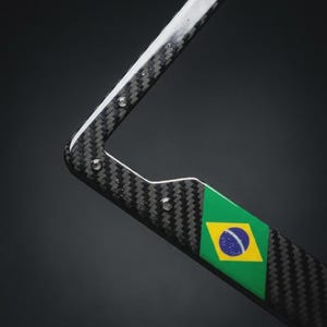 Puede incluir: Un marco para matrícula con un patrón de fibra de carbono en negro y gris. El marco incluye una sección rectangular que muestra la bandera de Brasil en verde, amarillo y azul. La estructura tiene orificios de montaje plateados visibles.