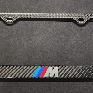 May include: A rectangular license plate frame featuring a black and grey carbon fiber pattern. The bottom center of the frame displays the letter M in white, preceded by three slanted stripes in light blue, dark blue, and red, representing the M logo.