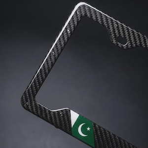 May include: A rectangular license plate frame featuring a black and gray carbon fiber weave pattern. The bottom section displays a green and white flag emblem with a crescent moon and a five-pointed star centered on the green background.