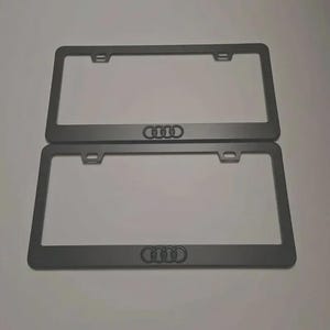 May include: Two rectangular matte black license plate frames featuring the Audi four-ring logo centered at the bottom. Each frame has two mounting holes at the top with horizontal slots for secure attachment to a vehicle's exterior bumper.