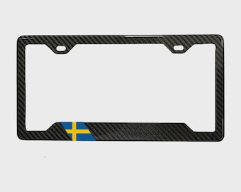 May include: A rectangular license plate frame featuring a dark gray carbon fiber pattern. The frame includes two mounting holes at the top and displays a small, rectangular Swedish flag emblem in blue and yellow on the bottom left corner.