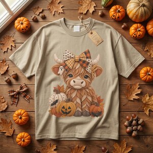 Op de afbeelding: Een beige t-shirt met korte mouwen en een afbeelding van een pluizige hooglandkoe met een geruite strik, omringd door pompoenen en herfstbladeren. De koe heeft kleine hoorns en een spookje. Een bruin papieren label zit aan de kraag vast.