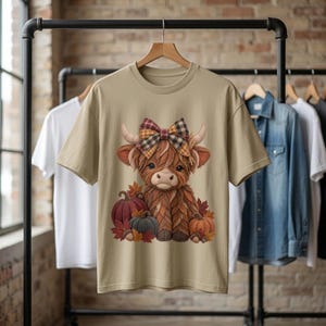 Op de afbeelding: Een beige t-shirt met korte mouwen en een afbeelding van een pluizige bruine hooglandkoe met een geruite strik. De koe is omringd door pompoenen en herfstbladeren in tinten oranje, rood en grijs, gecentreerd op de voorzijde van het kledingstuk.
