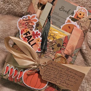 May include: A gift-wrapped book featuring autumn-themed paper with pumpkin and leaf illustrations. It includes a Hello Fall sticker, a gold-toned pen, a black tassel, a tea bag, a bookmark, and a handwritten tag labeled Autumn Romance.