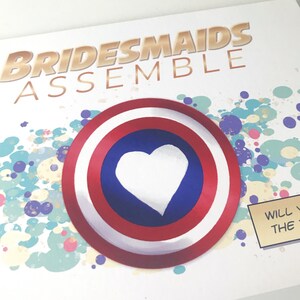 May include: A white card with the text "Bridesmaids Assemble" in gold letters. A red, white, and blue circle with a white heart in the center is on the card. The background is a colorful abstract design.
