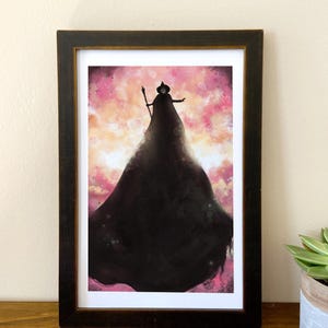 May include: Framed art print featuring a silhouetted figure in a long black cloak and hat, holding a staff. The background is a watercolor-style wash of pink, orange, and white. The frame is dark brown.