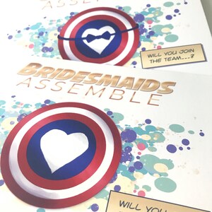 May include: Two invitations with a superhero theme. The invitations are white with gold text and a red, white, and blue shield with a white heart in the center. The text on the invitations reads "Groomsmen Assemble" and "Bridesmaids Assemble". The invitations also include the text "Will you join the team...?"