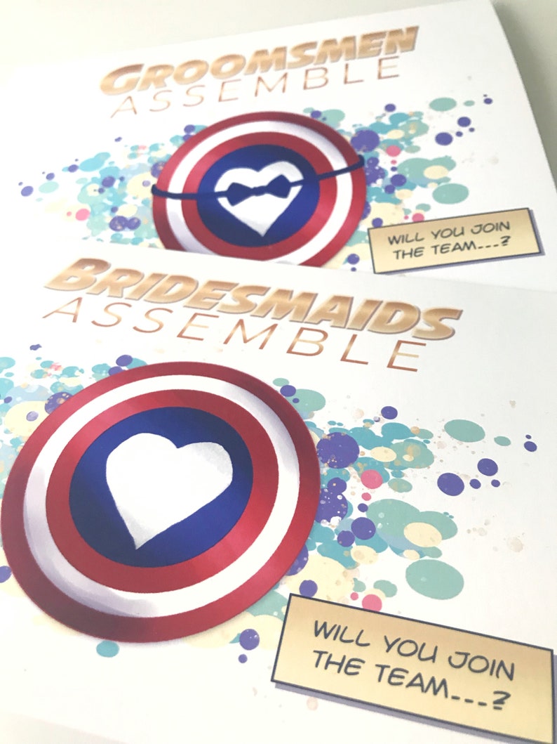 May include: Two invitations with a superhero theme. The invitations are white with gold text and a red, white, and blue shield with a white heart in the center. The text on the invitations reads "Groomsmen Assemble" and "Bridesmaids Assemble". The invitations also include the text "Will you join the team...?"