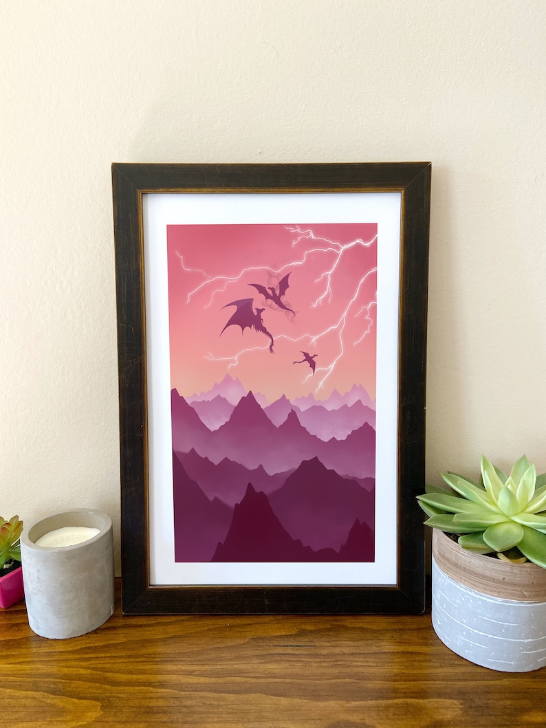 May include: A framed print of a digital illustration depicting three dragons flying through a pink and purple mountain range with lightning strikes in the sky.