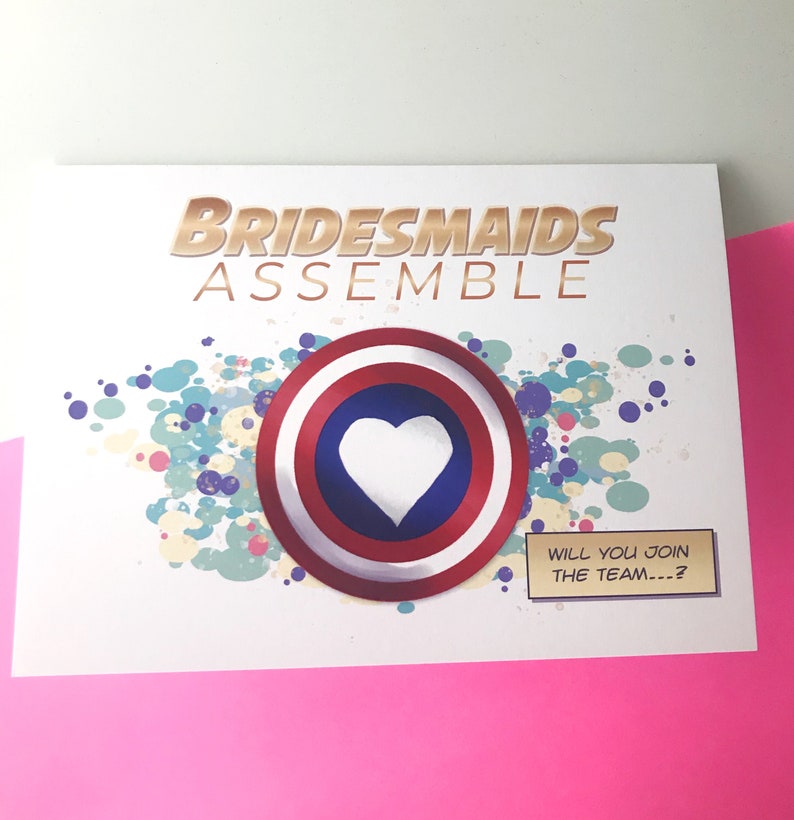 May include: A white card with the text "Bridesmaids Assemble" in gold lettering. A red, white, and blue circle with a white heart in the center is on the card. The text "Will you join the team...?" is in a comic book style speech bubble.