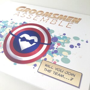 May include: A white card with a gold and red shield with a white heart and a black bow tie. The background is a colorful splatter pattern. The text "Groomsmen Assemble" is at the top of the card. The text "Will you join the team...?" is in a gold box with a black border.