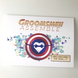 May include: A white card with the text "Groomsmen Assemble" in gold letters. A red, white, and blue shield with a white heart and a black bowtie is in the center of the card. The text "Will you join the team...?" is in a white speech bubble.