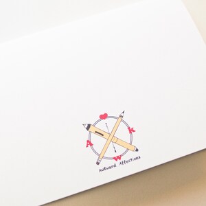 May include: White card with a compass design featuring two pencils crossed over each other. The compass has the letters "AWK" in red and the text "Awkward Affections" below the compass.