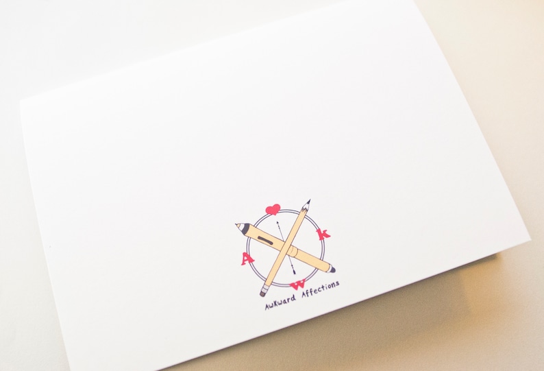 May include: White card with a compass design featuring two pencils crossed over each other. The compass has the letters "AWK" in red and the text "Awkward Affections" below the compass.