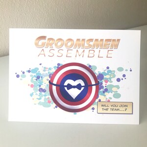 May include: A white card with the text "Groomsmen Assemble" in gold letters. A red, white, and blue shield with a white heart and a black bowtie is in the center of the card. The text "Will you join the team...?" is in a white box at the bottom of the card.
