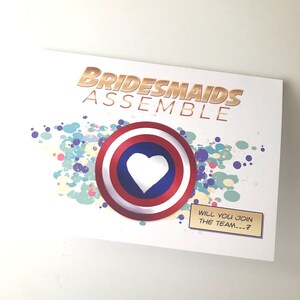 May include: A white card with the text "Bridesmaids Assemble" in gold letters. A red, white, and blue shield with a white heart in the center is on the card. The text "Will you join the team...?" is on a small brown rectangle.