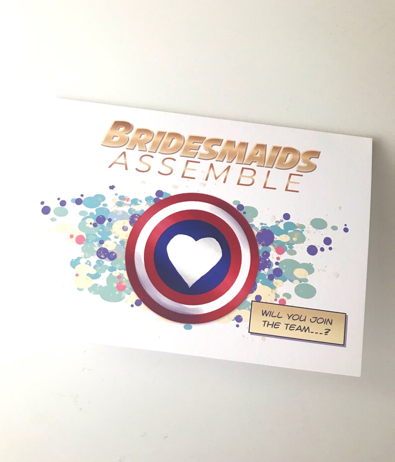 May include: A white card with the text "Bridesmaids Assemble" in gold letters. A red, white, and blue shield with a white heart in the center is on the card. The text "Will you join the team...?" is on a small brown rectangle.