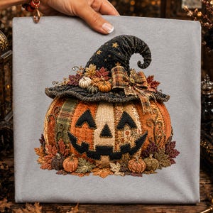 May include: A light gray t-shirt featuring a graphic of a patchwork jack-o'-lantern wearing a black witch hat with gold stars. The pumpkin is decorated with plaid fabric, autumn leaves, and small pumpkins in orange, brown, and green tones.