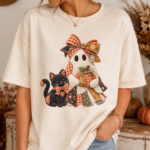 May include: A cream-colored short-sleeve t-shirt featuring a graphic of a ghost and a black cat. The ghost wears a large plaid bow and holds a pumpkin. The cat has a small orange bow. Both figures are designed with a patchwork quilt aesthetic.