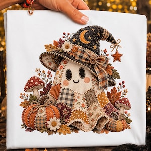 May include: A white t-shirt featuring a graphic of a patchwork ghost wearing a witch hat decorated with a crescent moon, stars, and a plaid bow. The ghost is surrounded by autumn leaves, two pumpkins, and two mushrooms in warm fall colours.