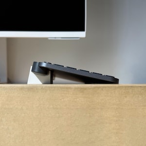 May include: A dark gray computer keyboard is positioned at an angle, resting on a white triangular stand. The keyboard features a cylindrical battery compartment on the left side and is set against a plain, light-colored background.
