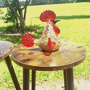 May include: A glass rooster figurine featuring a clear body with multicolored confetti-like speckles in blue, yellow, and red. The rooster has a solid red comb, a red wattle, a red tail, and a black beak, all crafted from smooth, polished glass.