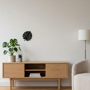Könnte beinhalten: Ein schwarzes Wandkunstobjekt aus Metall in Form eines Monstera-Blattes, das an einer schlichten weißen Wand über einem Medienboard aus hellem Eichenholz angebracht ist. Das Board hat zwei geschlossene Schränke und ein offenes Fach.