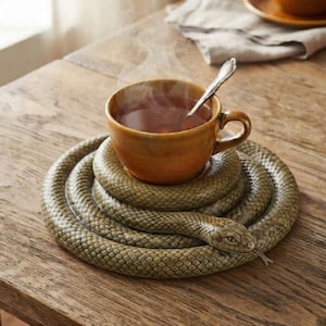 Könnte beinhalten: Eine braune Keramiktasse mit einem dampfenden Heißgetränk und einem Silberlöffel, die auf einem runden, schlangenförmigen Untersetzer steht. Die Schlangenfigur hat eine texturierte, schuppige Oberfläche in Olivgrün und Braun mit einem Kopf.