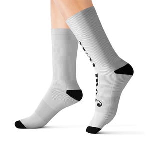 Puede incluir: Un par de calcetines blancos de corte medio con textura acanalada y detalles negros en talones y punteras. El texto "STAY COOL" está impreso verticalmente en una fuente negra, negrita y estilizada a lo largo del lateral de la tela.