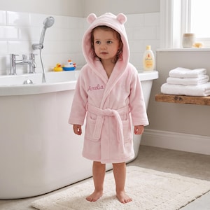 May include: A soft, light pink hooded bathrobe featuring two small rounded ears on the hood and a tie-waist belt. The front of the robe is embroidered with the name Amelia in a cursive script, and it includes two front patch pockets.
