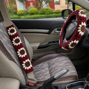 Könnte beinhalten: Ein gehäkelter Lenkradbezug und ein passender Sicherheitsgurtbezug mit einem wiederkehrenden Muster aus dunkelroten Quadraten mit weißen und braunen Blumenmotiven. Die Accessoires sind an einem Autolenkrad und einem beigen Sicherheitsgurt angebracht.