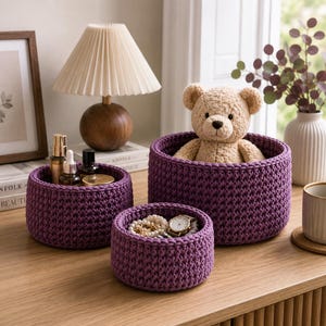May include: A set of three round, deep purple crocheted storage baskets of varying sizes. The largest basket contains a beige plush teddy bear, the medium basket holds several small glass bottles, and the smallest basket contains jewelry.