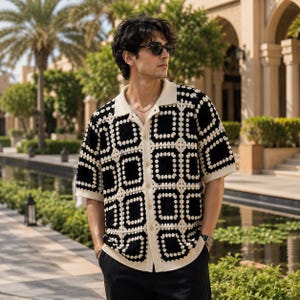 May include: A short-sleeved button-down shirt featuring a black and cream crochet pattern of square motifs. The shirt has a solid cream-colored collar and a front placket with matching buttons, constructed with a textured, open-knit fabric design.