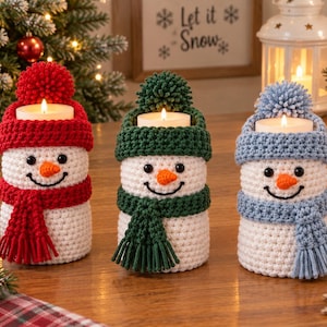 May include: Three cylindrical white crochet snowman candle holders, each topped with a beanie and matching scarf in red, dark green, or light blue. Each snowman features black bead eyes, an orange carrot nose, and a stitched black smile.